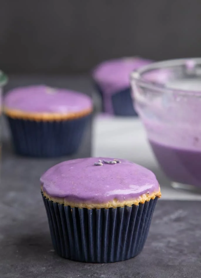 Yellow cupcake with purple glaze and lavender flower buds on gray surface.