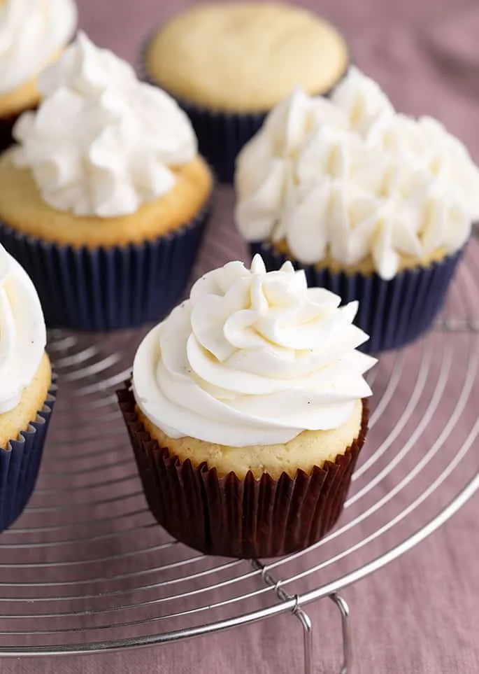 Yellow cupcakes with frosting in paper liners on round wire rack above purple cloth