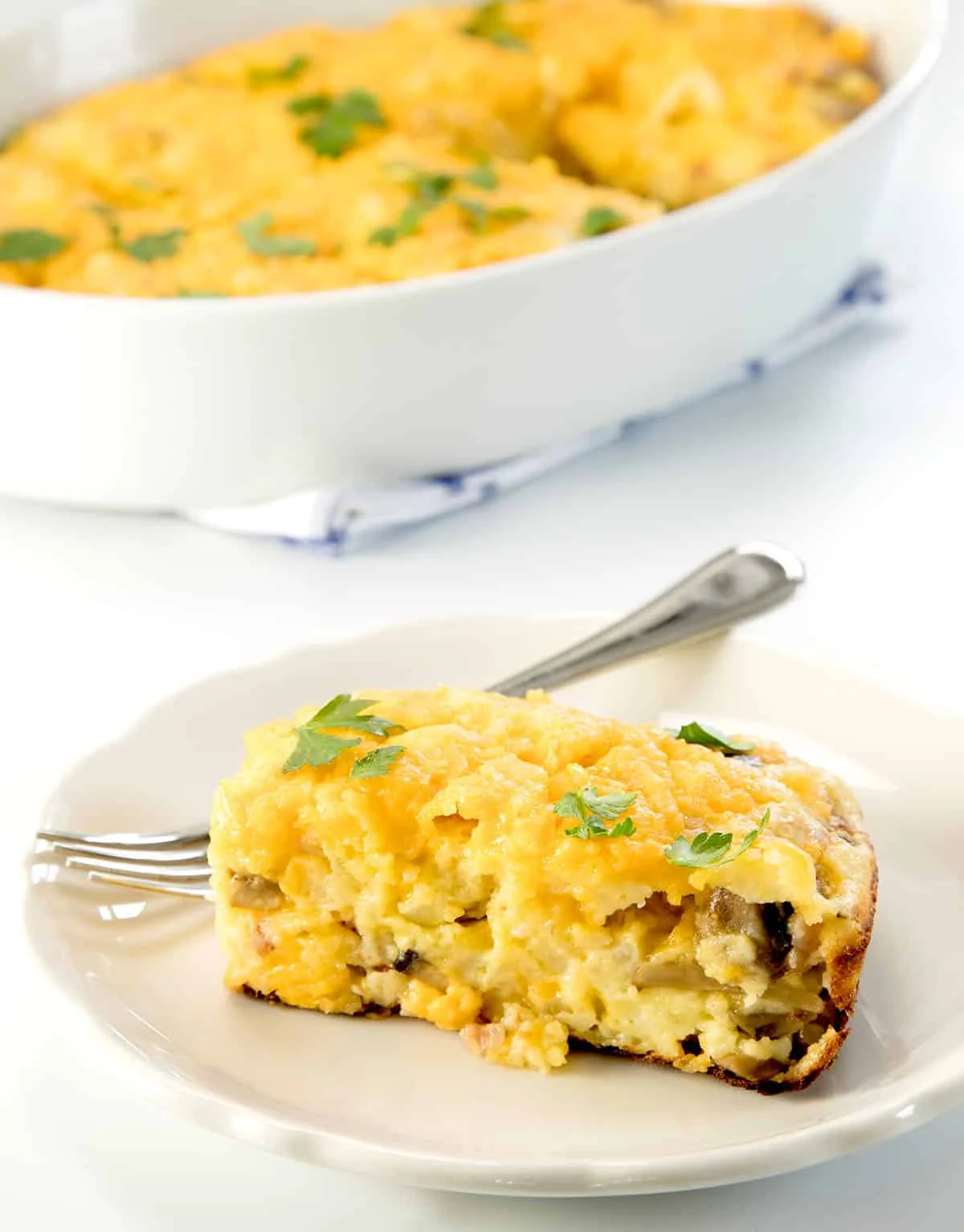 yellow rectangular slice of breakfast casserole with mushrooms inside and green parsley leaves on top on small white plate with small metal fork