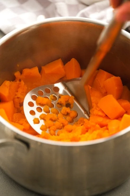 A potato masher being used to mash cooked sweet potatoes in a pot.