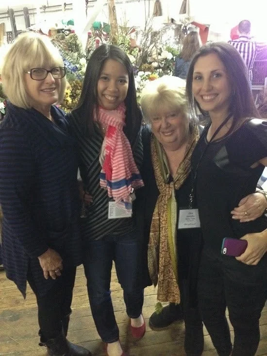 Four women smiling with their arms around each other and a flower arrangement in the background