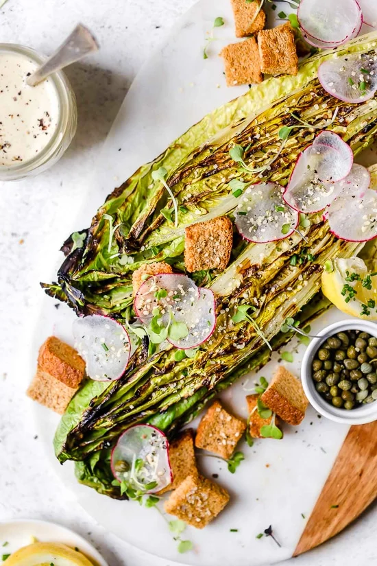 Grilled romaine salad with vegan Caesar dressing on marble and wood board.