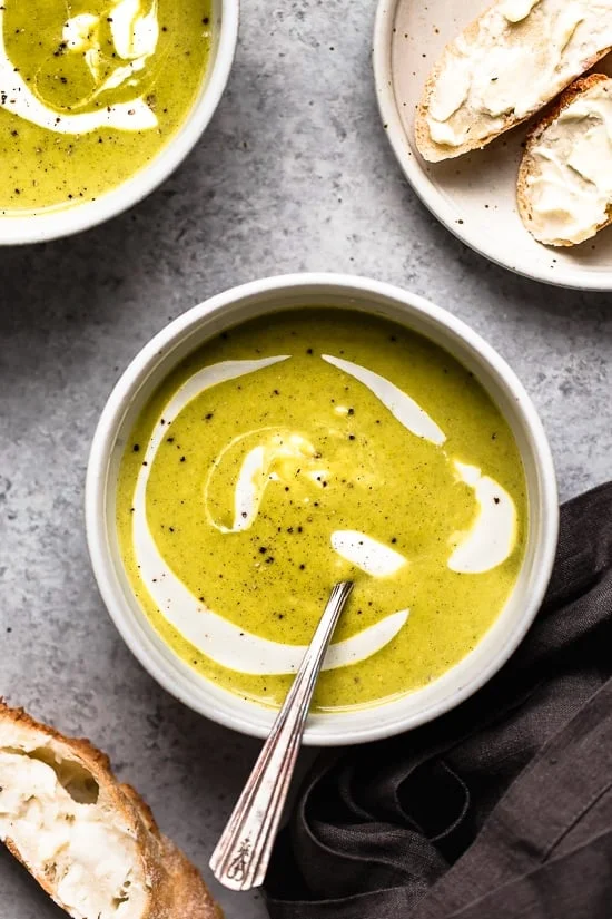 Overhead view of a bowl of homemade creamy broccoli soup drizzled with sour cream next to crusty bread slices.