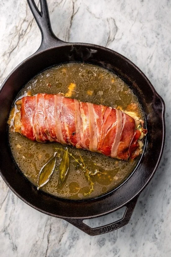 Skillet with prosciutto-wrapped stuffed turkey tenderloin and broth.